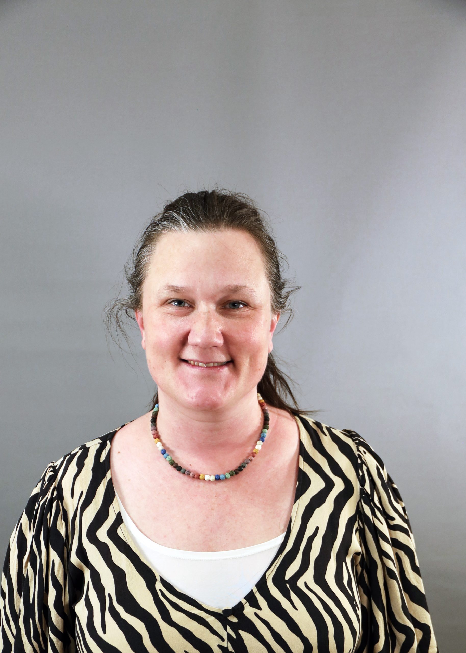 Leende kvinna med zebramönstrad topp och pärlhalsband, stående framför en enfärgad grå bakgrund.