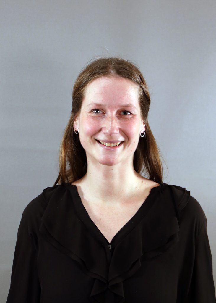 En kvinna med långt brunt hår och svart blus ler mot en grå bakgrund.