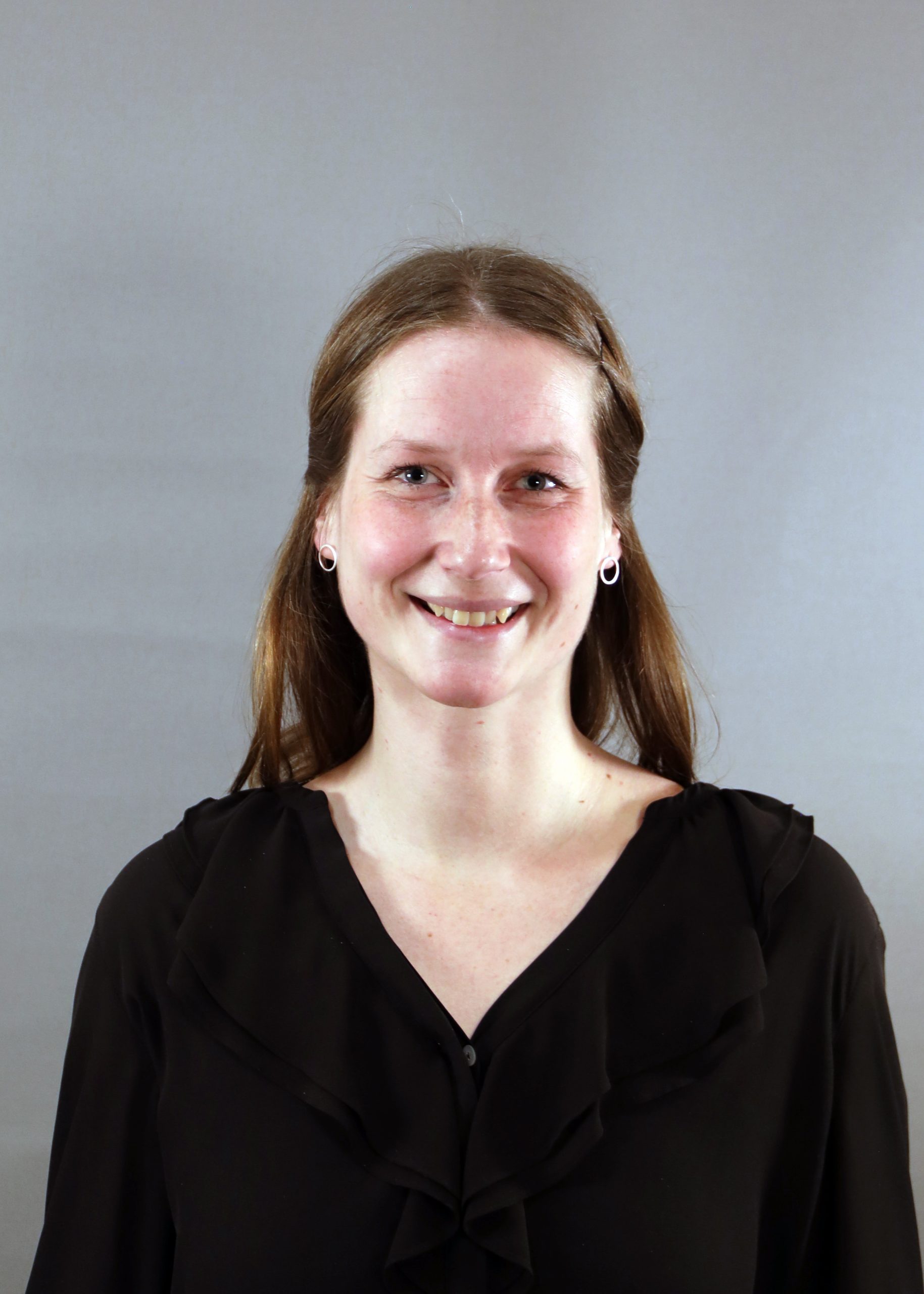 En kvinna med långt brunt hår och svart blus ler mot en grå bakgrund.