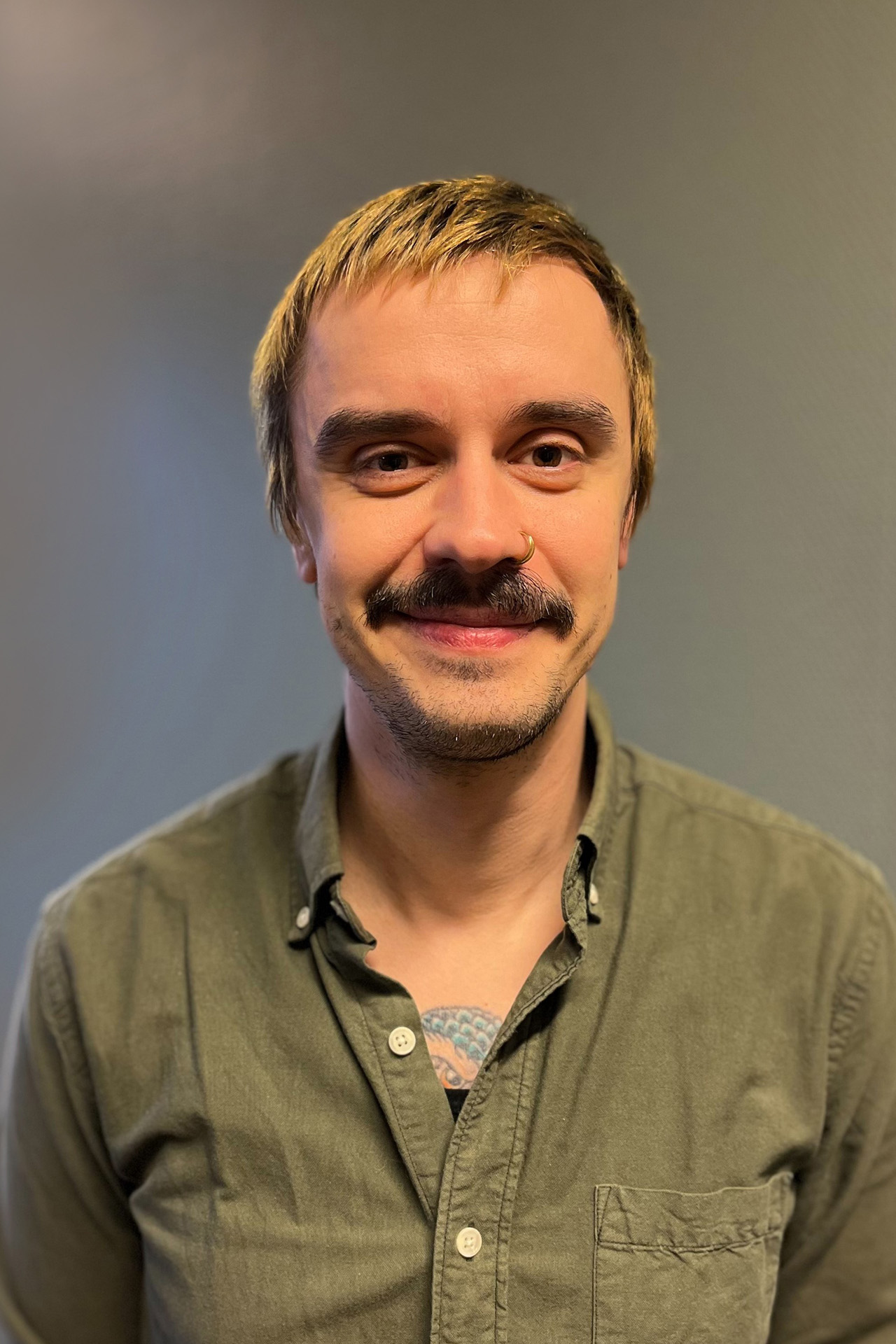 Leende person med mustasch, klädd i grön skjorta, står framför en enfärgad grå bakgrund.