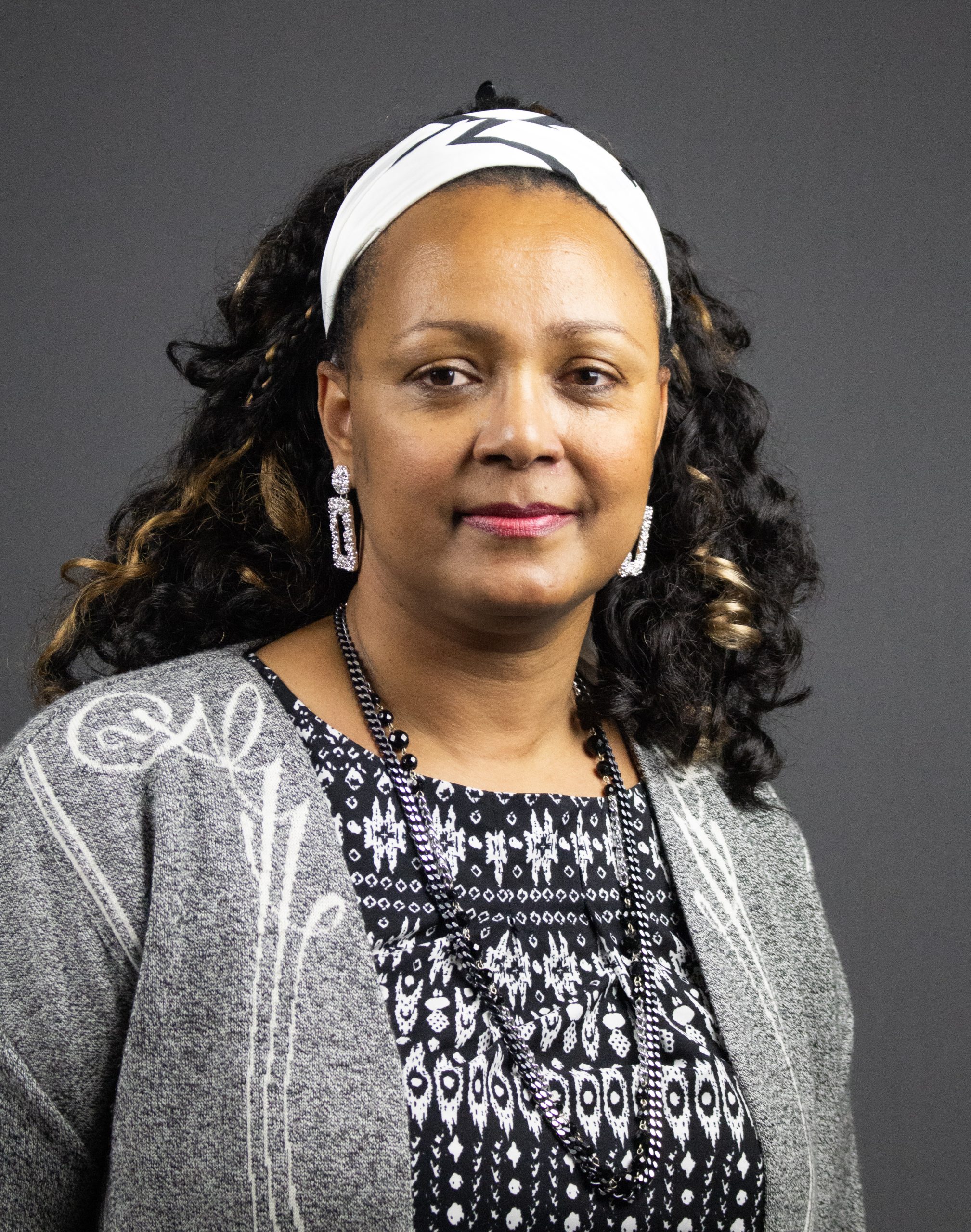 Kvinna med långt lockigt hår, iförd pannband, örhängen, mönstrad topp och grå kofta, mot grå bakgrund.