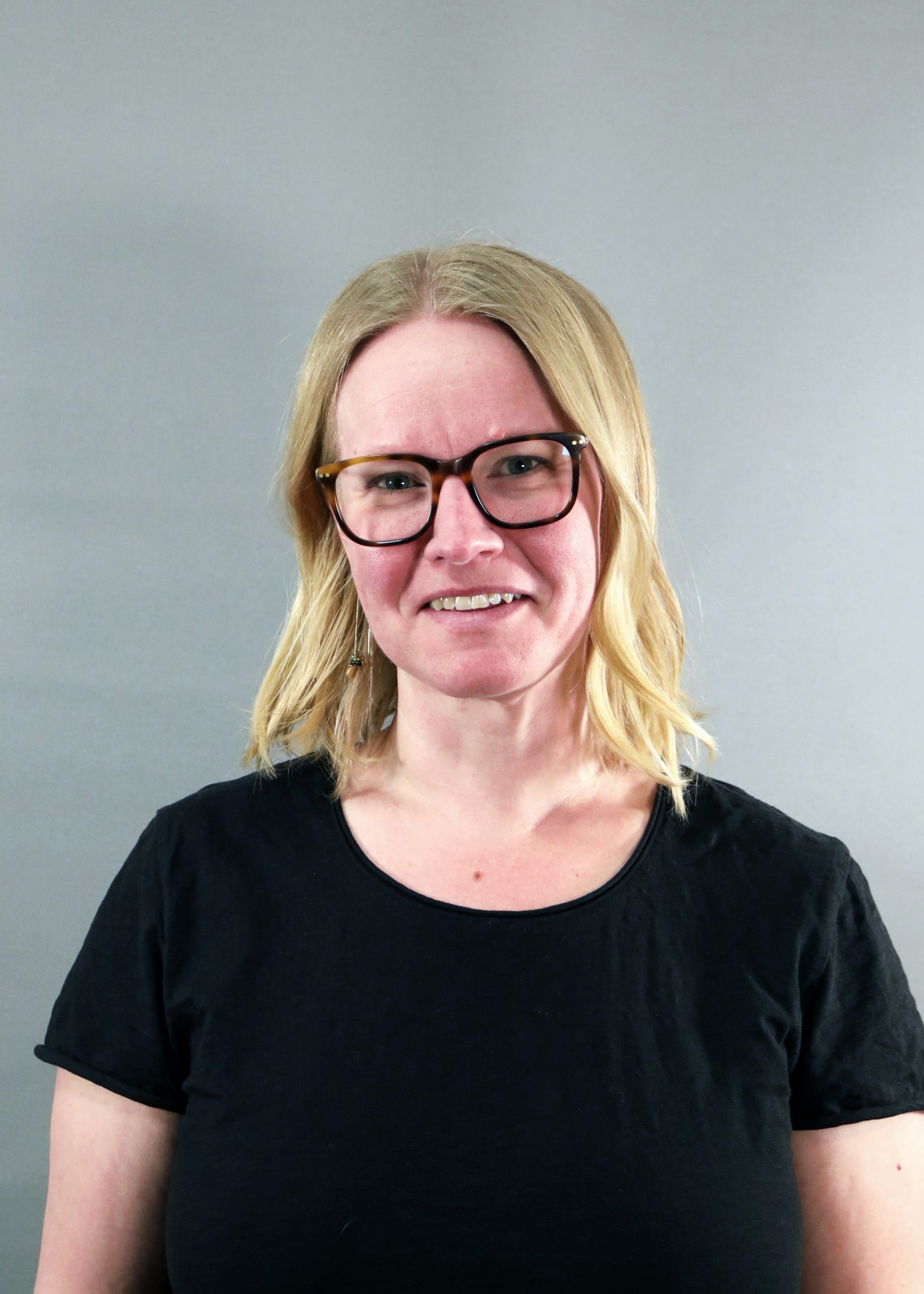 Kvinna med blont hår och glasögon i svart skjorta ler mot en grå bakgrund.