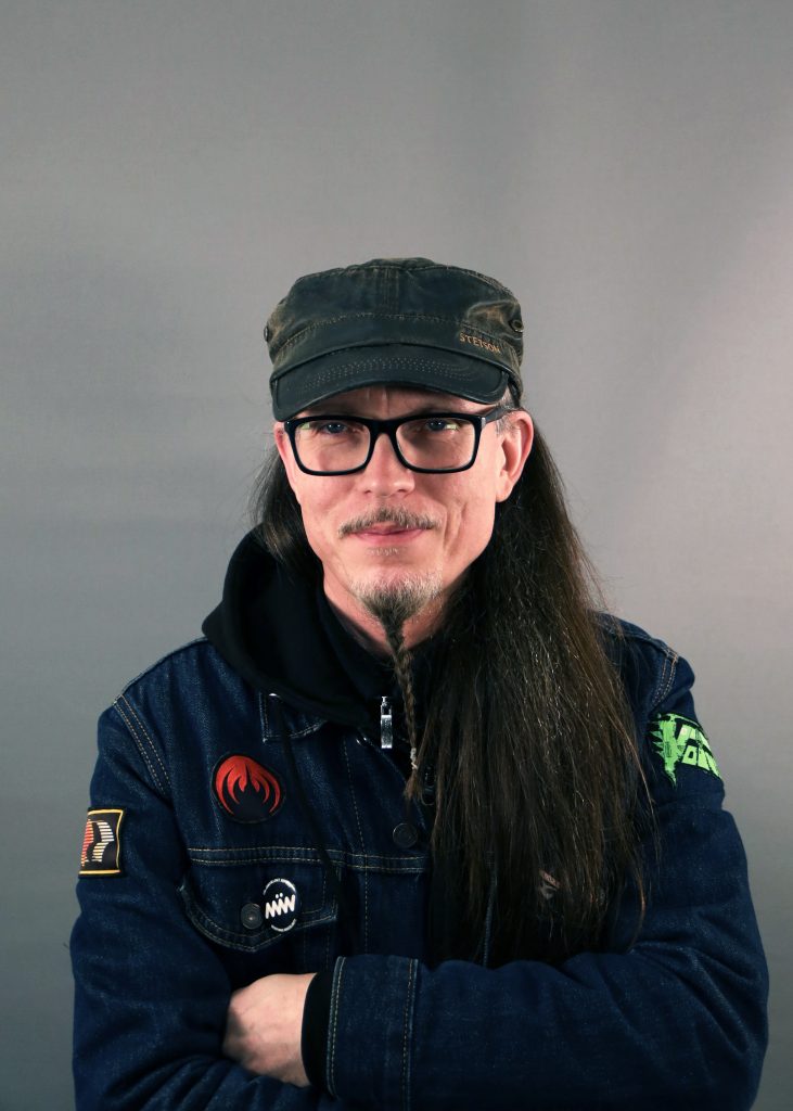 En person med långt hår, glasögon och keps iklädd jeansjacka står med armarna i kors mot en grå bakgrund.