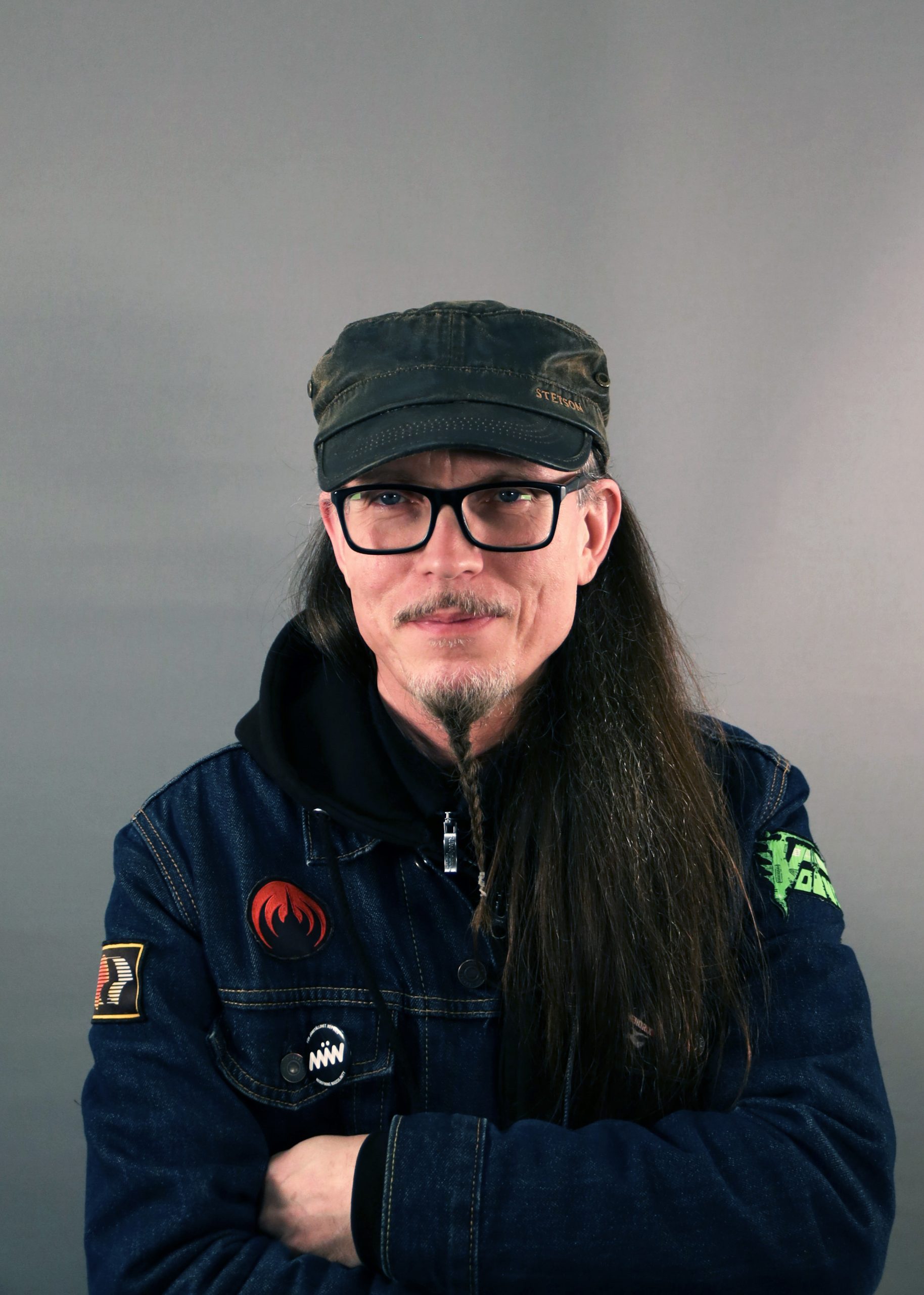 En person med långt hår, glasögon och keps iklädd jeansjacka står med armarna i kors mot en grå bakgrund.