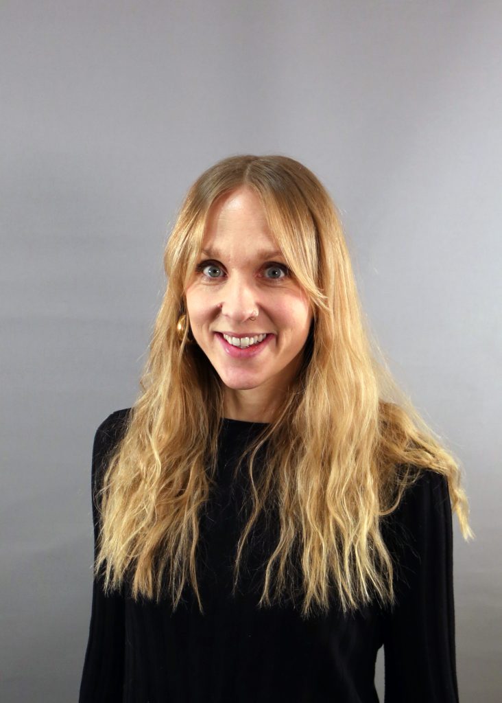 En kvinna med långt blont hår och svart topp ler mot en grå bakgrund.