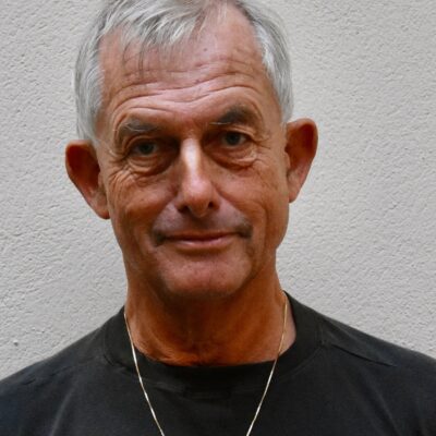 Äldre man med kort grått hår, iförd mörk skjorta och halsband, ler lätt mot en ljus vägg.