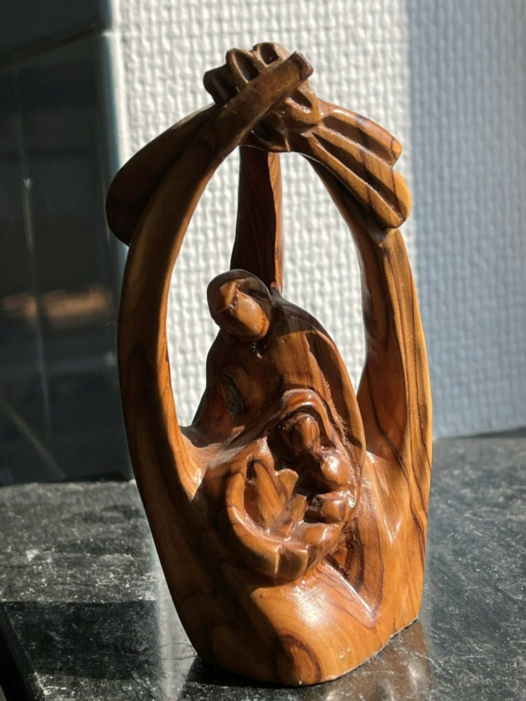 Träskulptur av två abstrakta figurer som omfamnar varandra med armarna över huvudet, på en mörk yta.