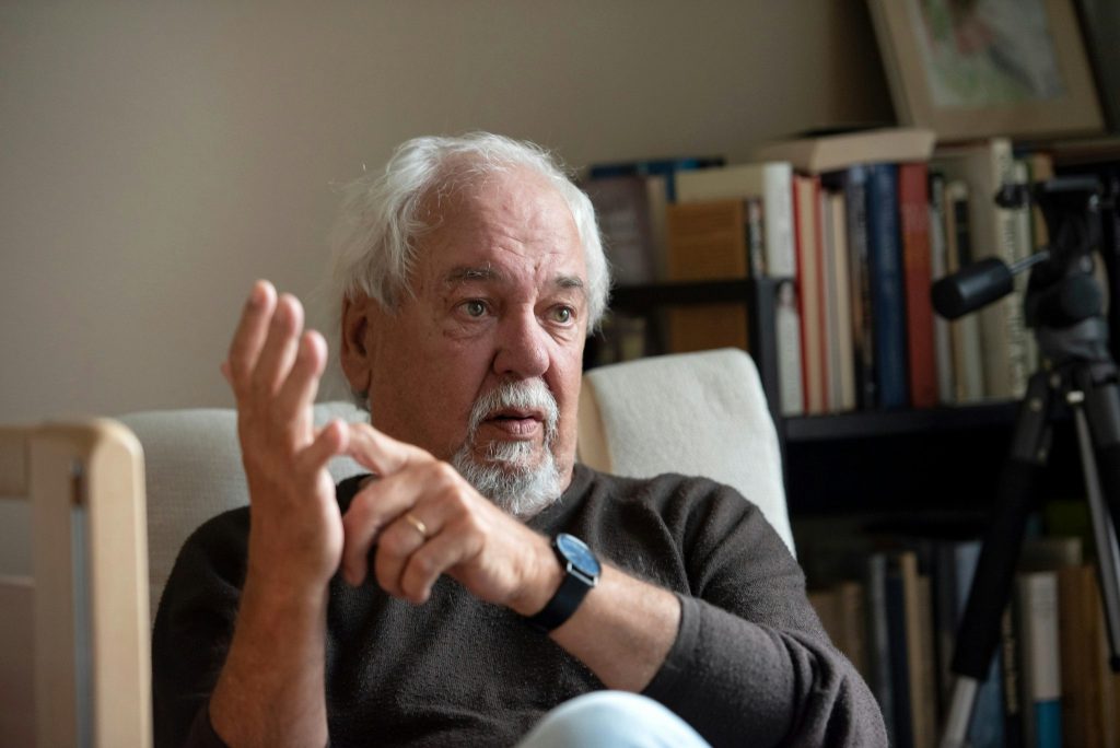 Äldre man med vitt hår och skägg gestikulerar medan han sitter i en stol med bokhyllor i bakgrunden.