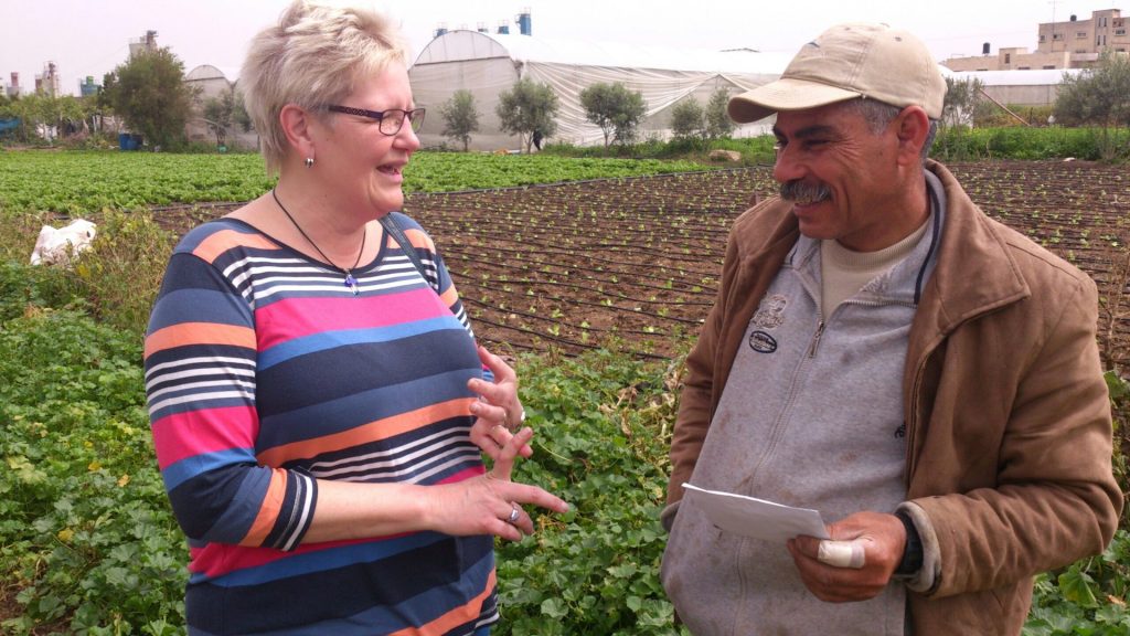 Två personer pratar och ler medan de står på ett grönt fält med grödor och ett växthus i bakgrunden.