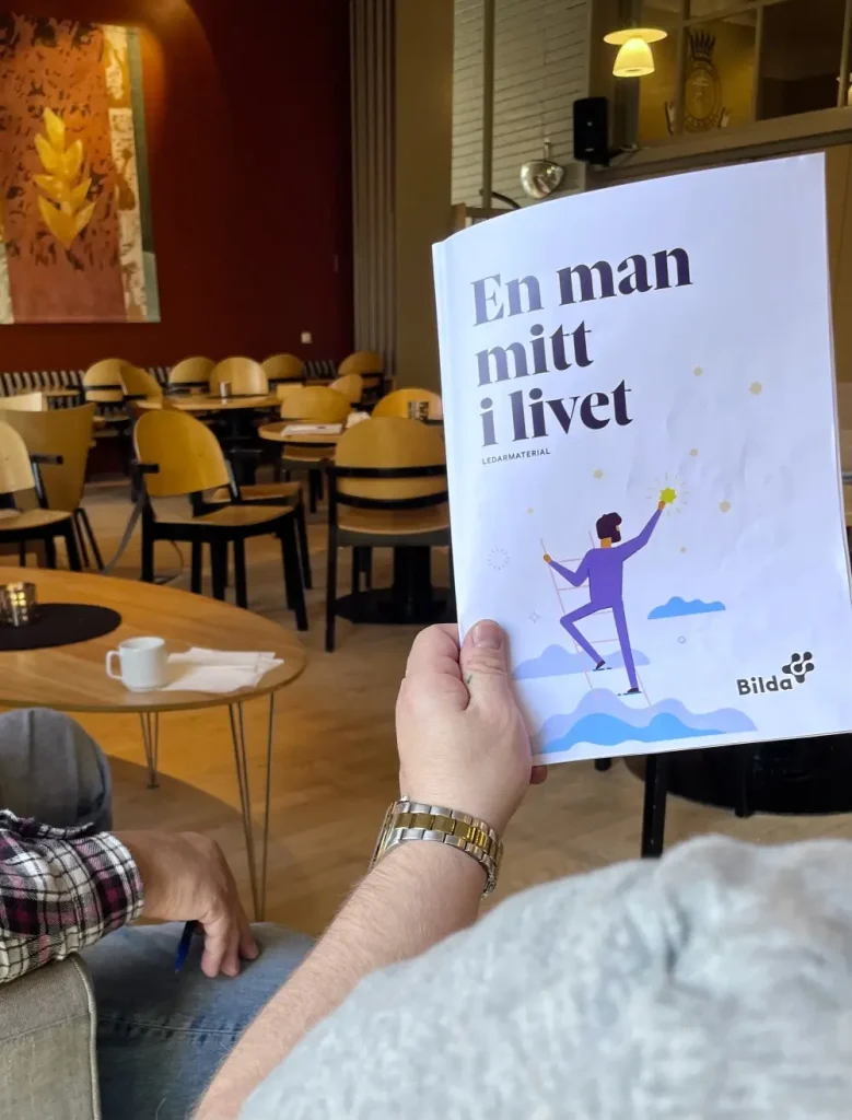 En person håller i en svensk broschyr med titeln "En man mitt i livet" på ett mysigt café med tomma stolar och bord.