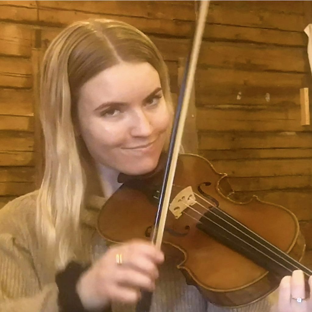 Kvinna med långt blont hår spelar fiol i ett mysigt trärum och ler mot kameran under en Hälsinge Låtverkstad-session.