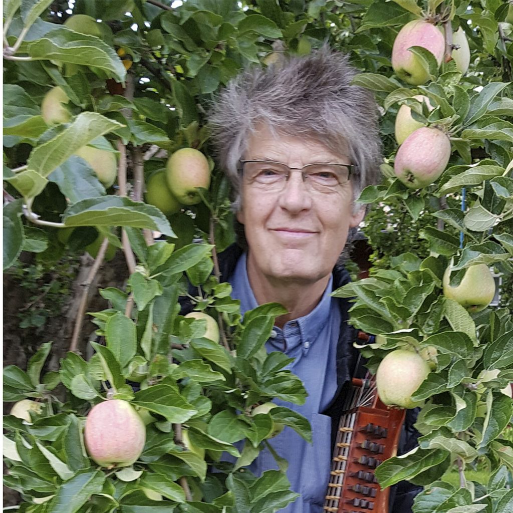 En äldre man med glasögon står bland äppelträdets grenar och håller i ett stränginstrument.