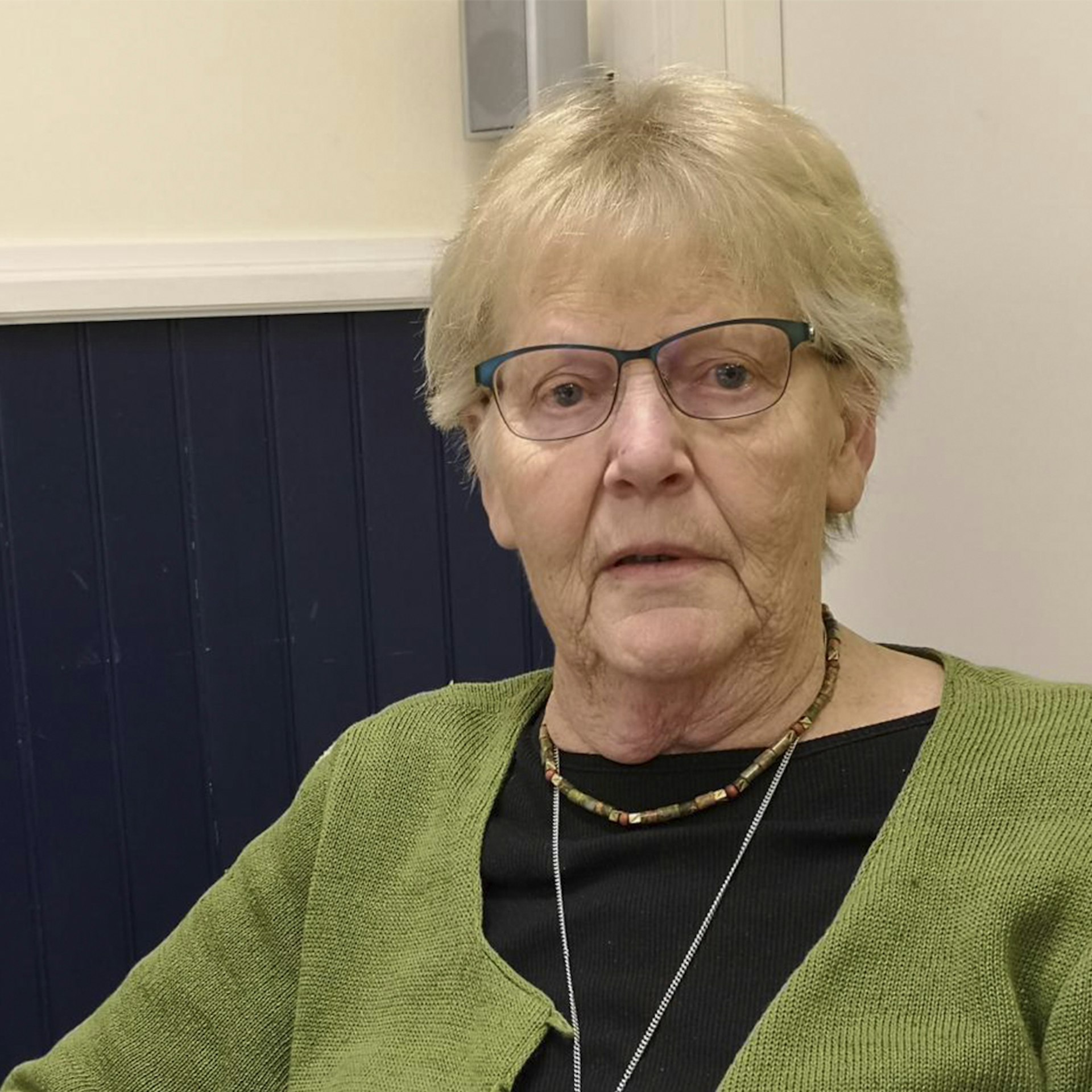 En äldre kvinna med glasögon och grön kofta sitter inomhus och tittar mot kameran.