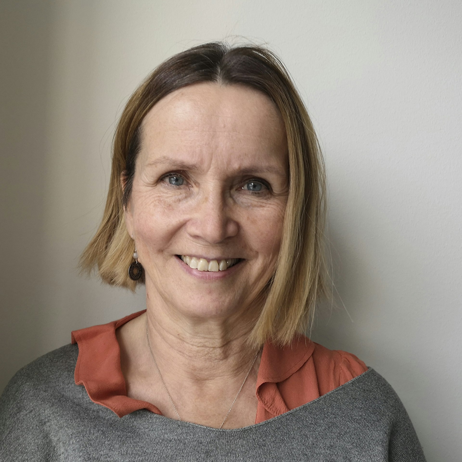 Leende kvinna med kort, rakt hår, klädd i grå topp och orange blus, mot ljus bakgrund.