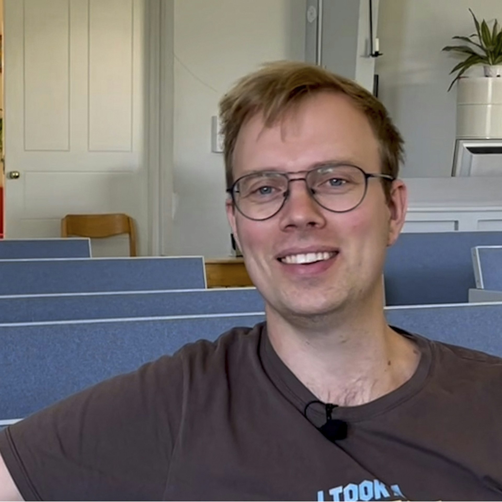 En leende man med glasögon sitter i ett klassrum med blåa stolar och en växt i bakgrunden.