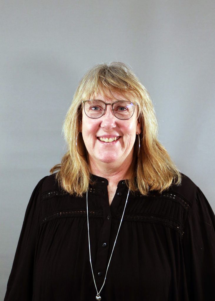 En leende kvinna med glasögon och blont hår, klädd i en svart topp och ett långt halsband, mot en grå bakgrund.