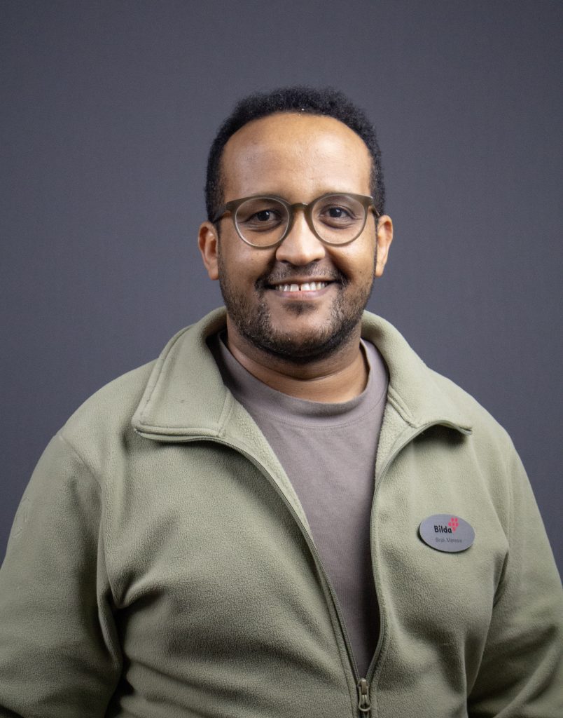 Man med glasögon och grön jacka ler mot en grå bakgrund.