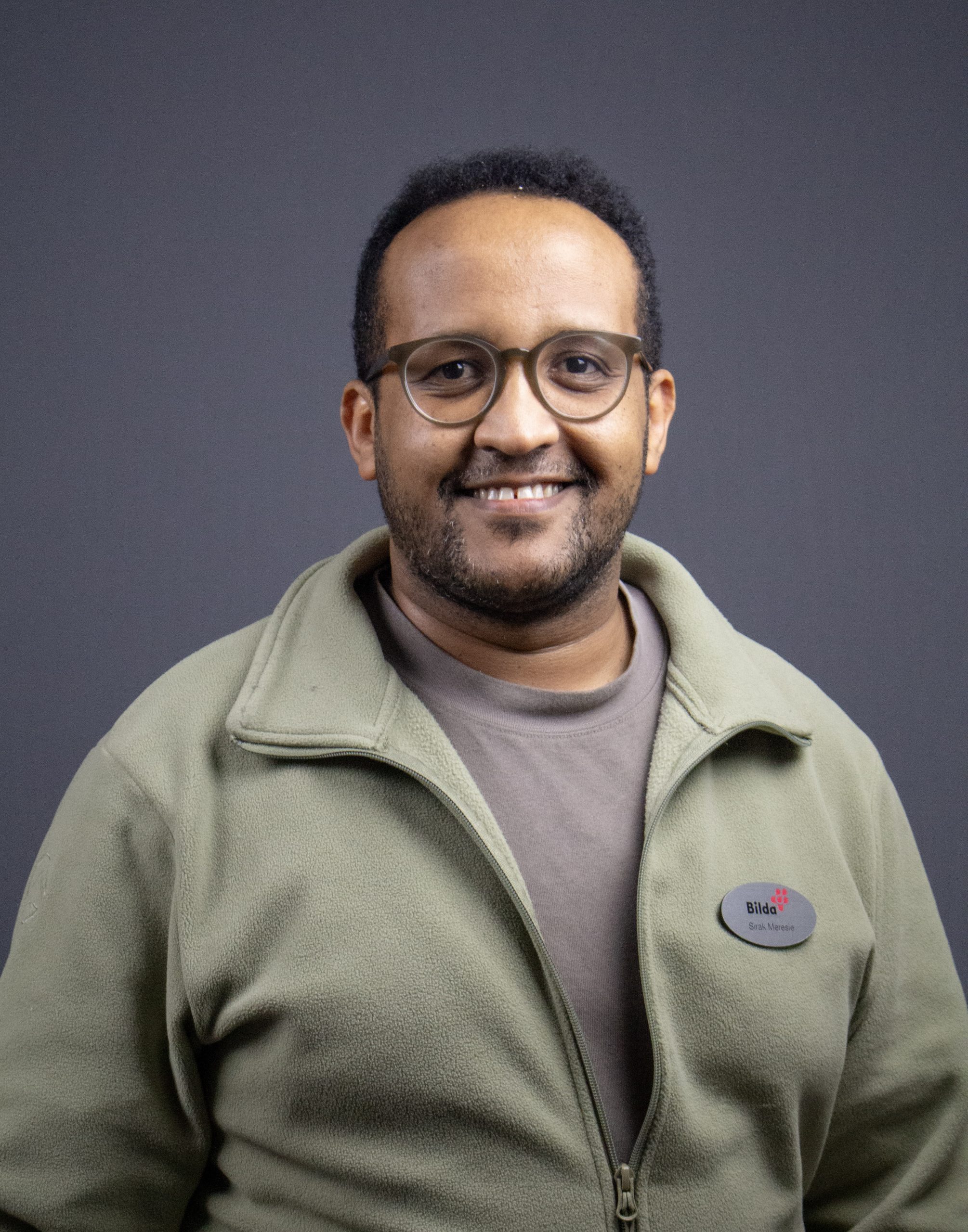 Man med glasögon och grön jacka ler mot en grå bakgrund.