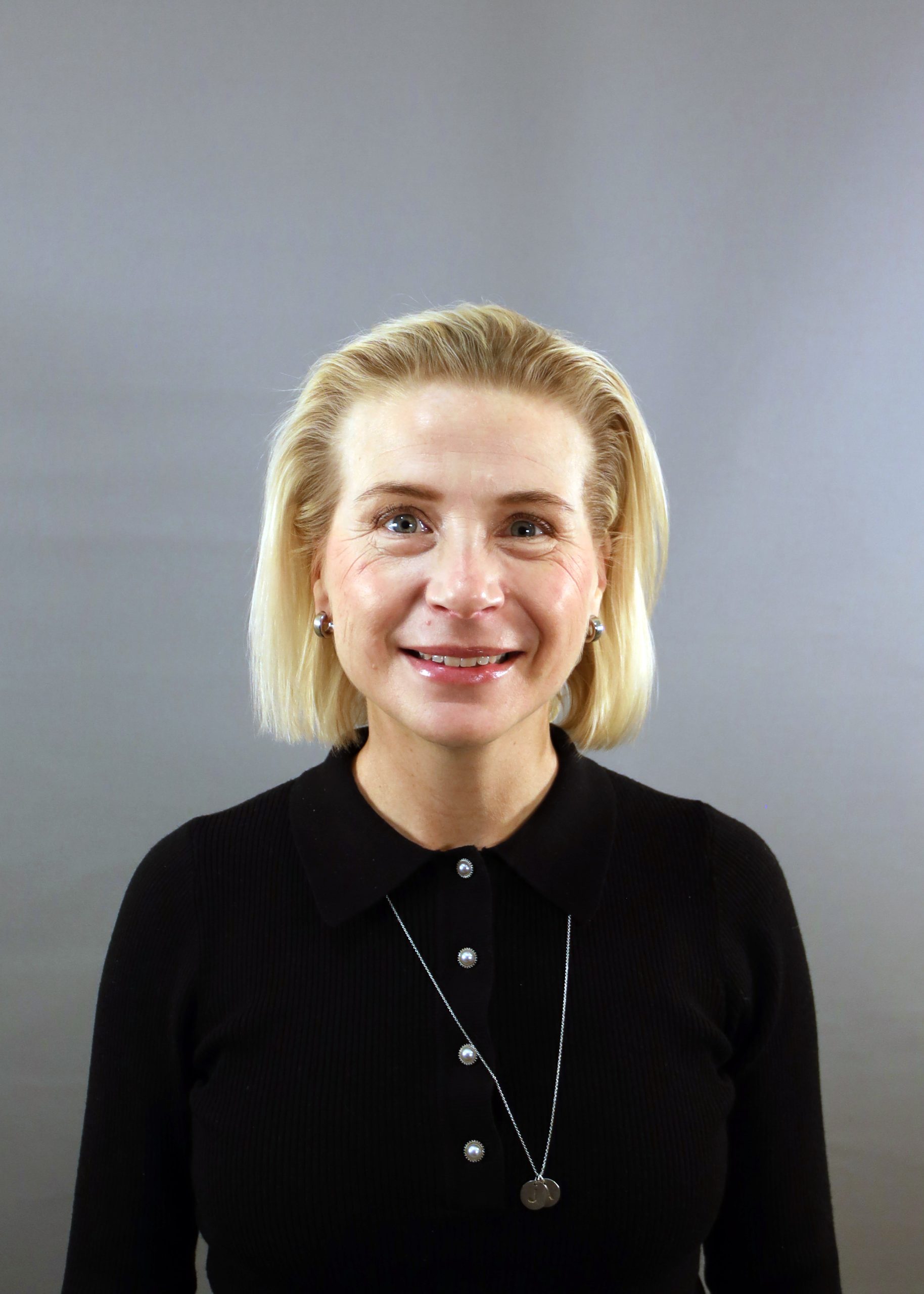 En kvinna med blont hår, svart topp och halsband ler mot en grå bakgrund.