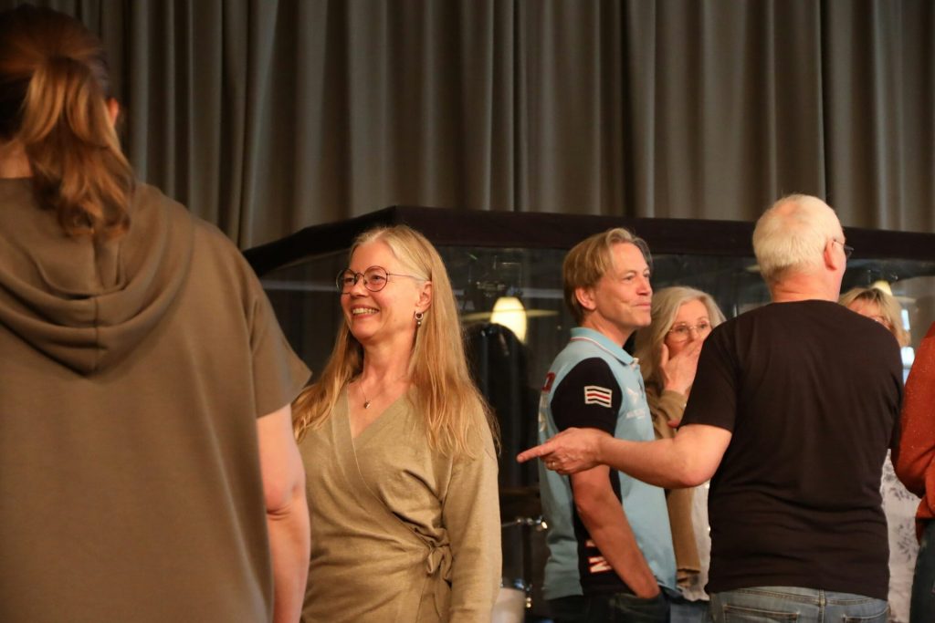 Människor står och pratar i en grupp inomhus, med en kvinna som ler i mitten.