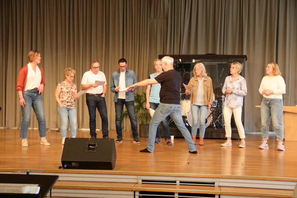 En grupp vuxna står på en scen och sjunger tillsammans, medan en man gestikulerar energiskt framför dem.