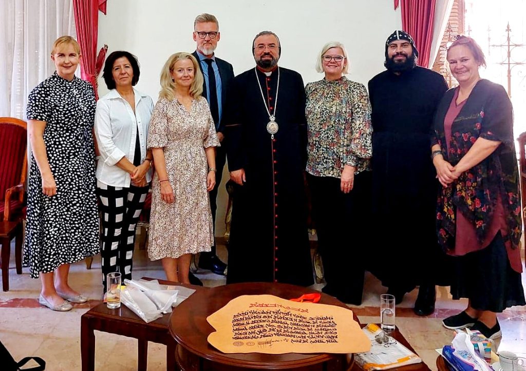 Sju personer står inomhus bredvid en sittande präst och på ett bord ligger ett stort orange blad med text.
