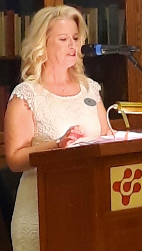 Kvinna i vit klänning talar vid ett podium med mikrofon i en biblioteksmiljö.