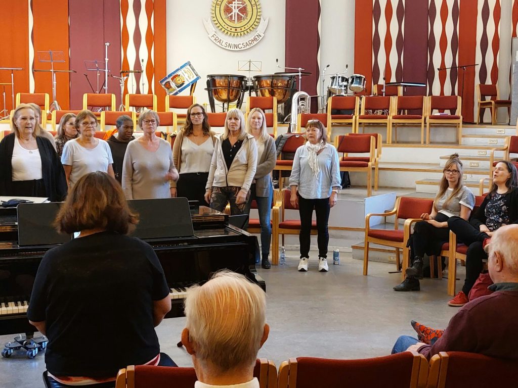 En kvinnokör sjunger medan en publik lyssnar i ett musikrum med instrument och röda stolar.