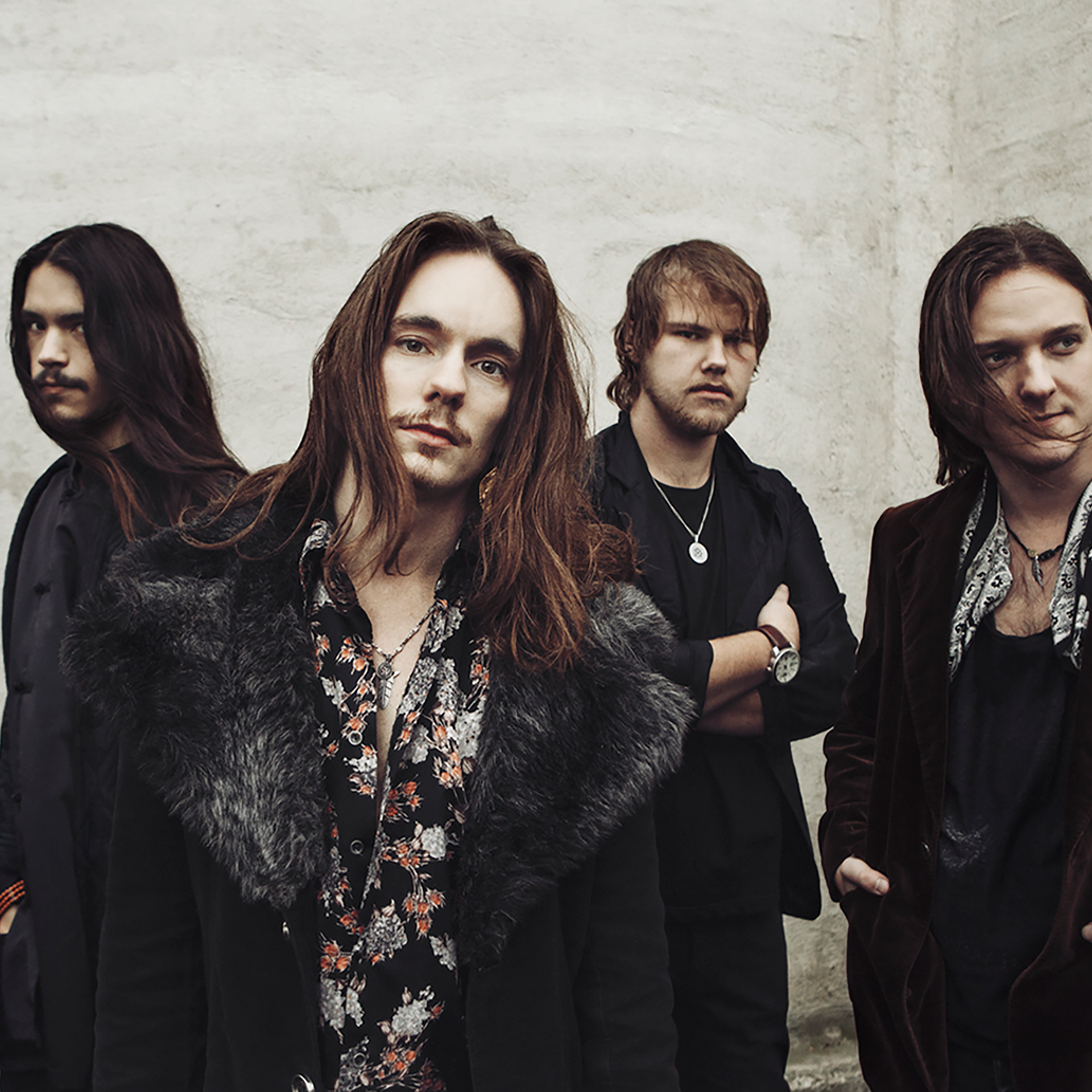 Fyra män med långt hår står mot en betongvägg, klädda i mörka, stilrena kläder och med allvarliga ansiktsuttryck.