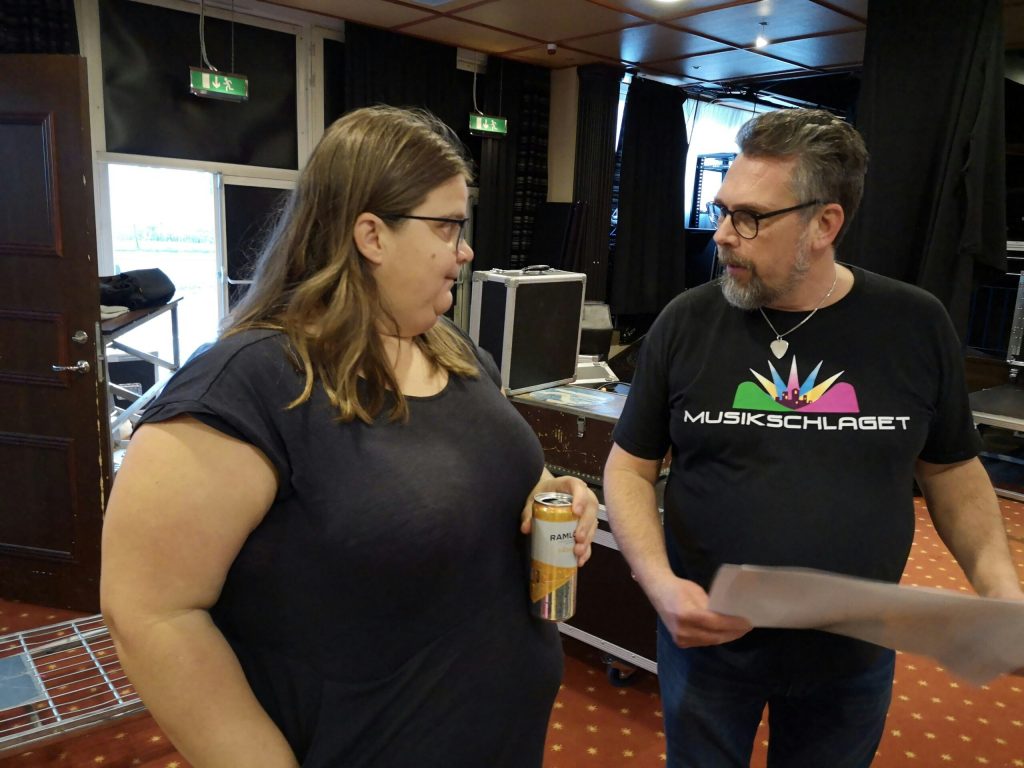 Två personer pratar inomhus; den ena håller i en drinkburk, den andra håller i papper; musikutrustning i bakgrunden.