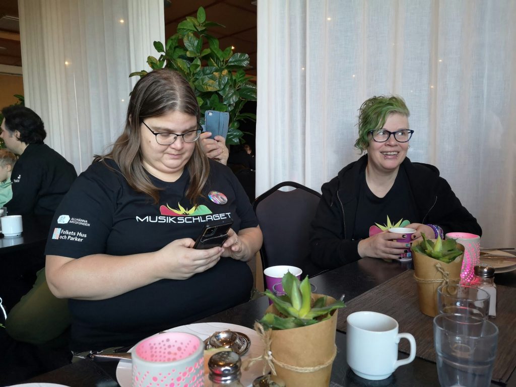 Två kvinnor sitter vid ett bord med drinkar och krukväxter, den ena använder en telefon, båda bär svarta skjortor.