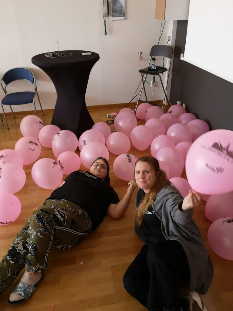 Två personer ligger på ett trägolv omgivet av många rosa ballonger i ett rum med stolar och bord.