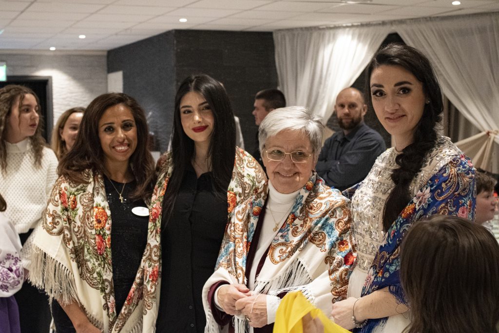 Fyra kvinnor i färgglada sjalar ler vid en sammankomst, med människor och draperade gardiner i bakgrunden.