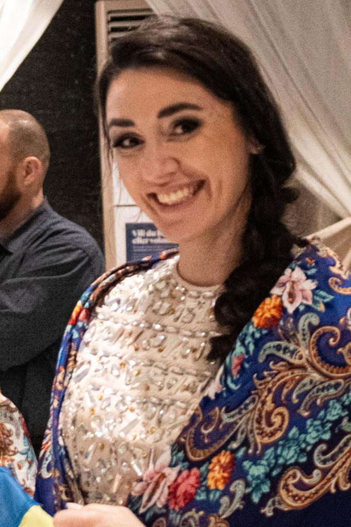 En kvinna med mörkt hår i en fläta, bär en mönstrad sjal och ler vid ett evenemang inomhus.