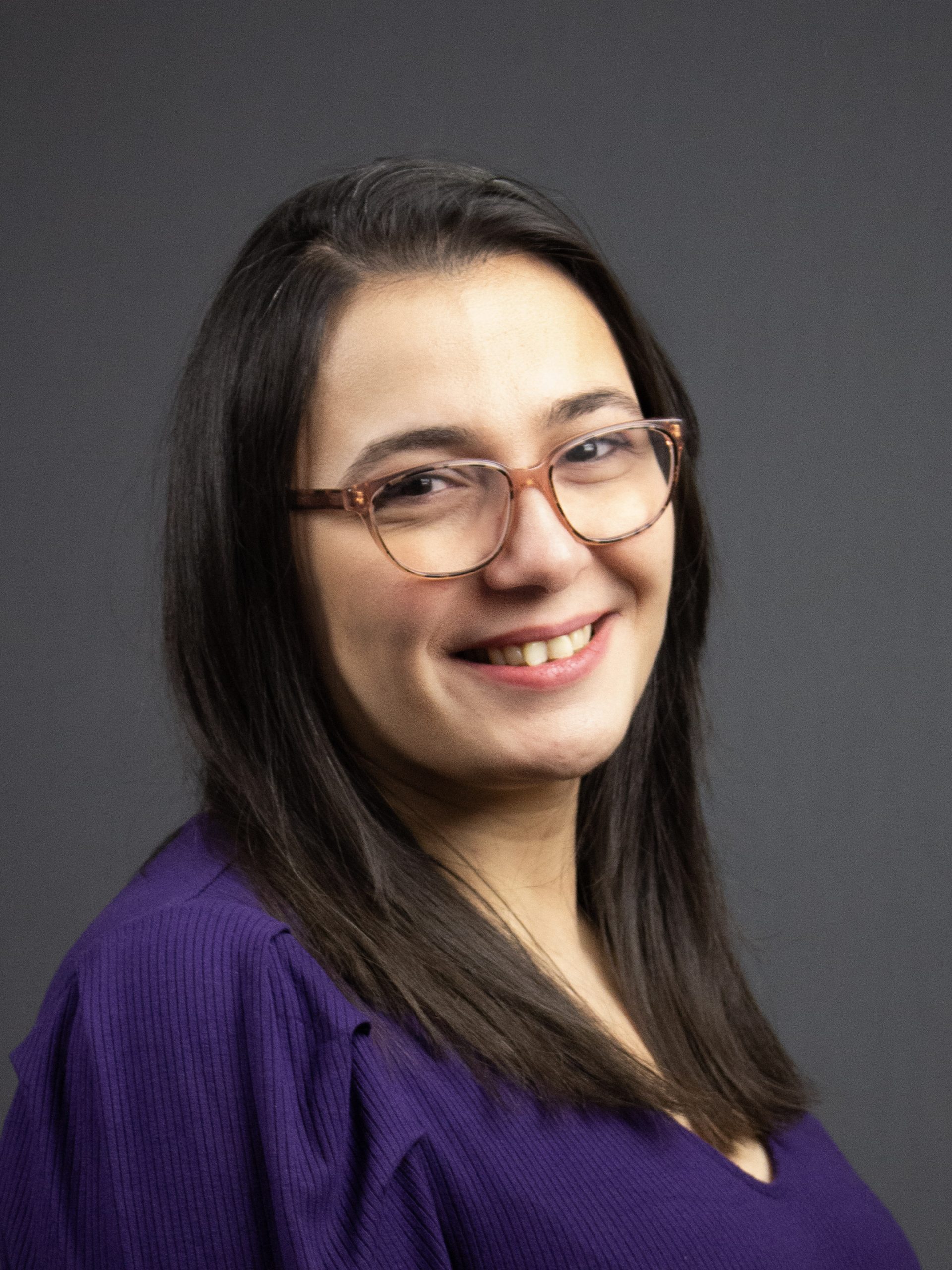 Kvinna med långt mörkt hår och glasögon ler, klädd i en lila topp mot en vanlig mörk bakgrund.