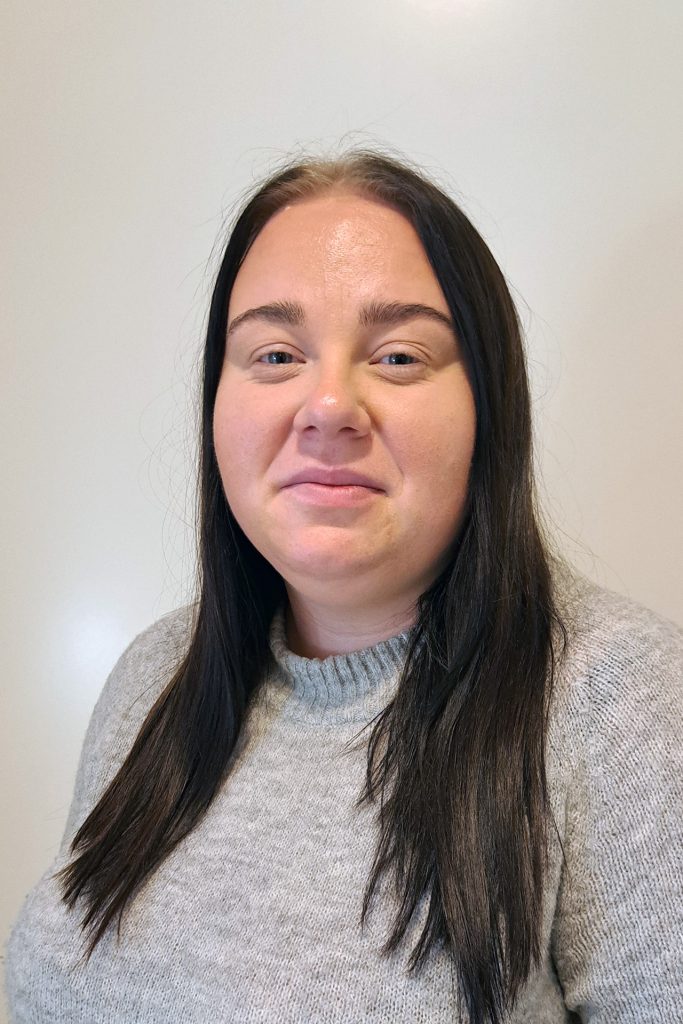 Kvinna med långt mörkt hår och grå tröja ler mot en ljus bakgrund.