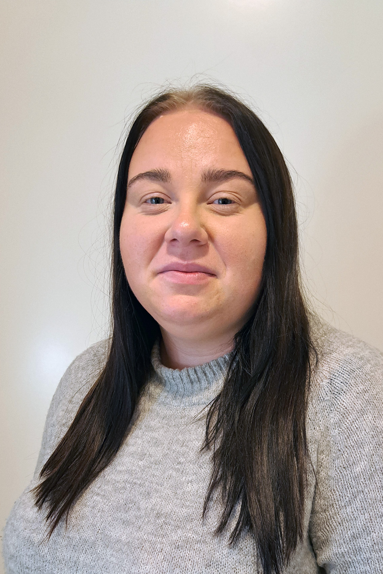 Kvinna med långt mörkt hår och grå tröja ler mot en ljus bakgrund.