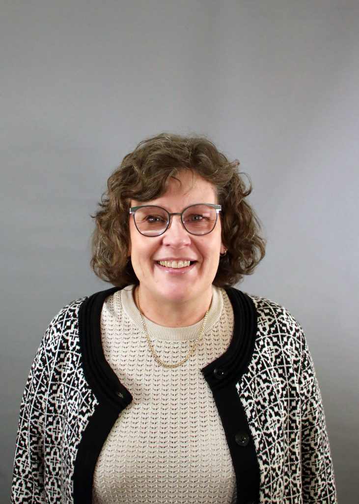 Kvinna med lockigt brunt hår, glasögon och en mönstrad tröja ler mot en grå bakgrund.