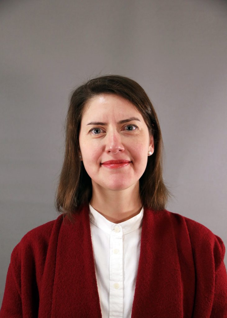 Kvinna med axellångt brunt hår i röd kappa och vit skjorta, stående mot grå bakgrund.