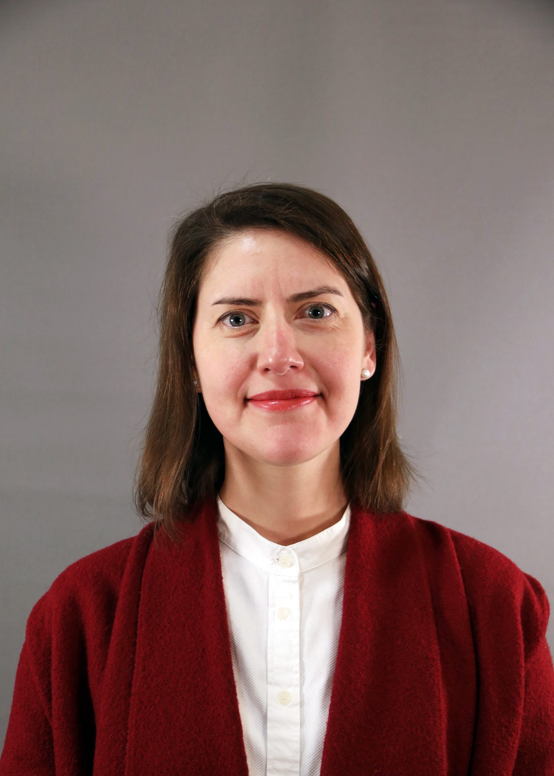Kvinna med axellångt brunt hår i röd kappa och vit skjorta, stående mot grå bakgrund.