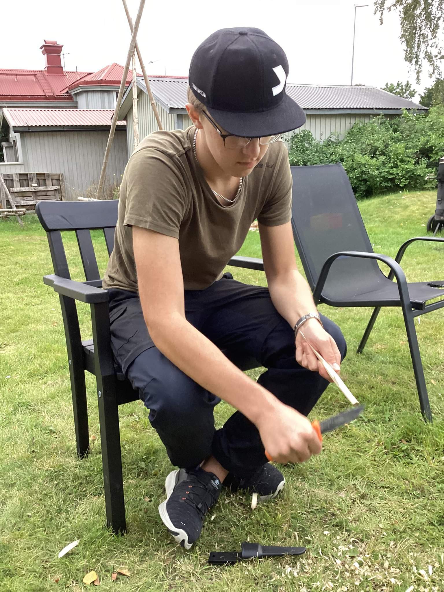 En scoutledare sitter på en svart utomhusstol och täljer en pinne med en kniv på en gräsmatta.