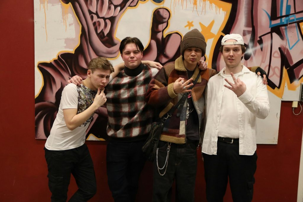 Fyra unga män poserar tillsammans framför färgglad graffiti och gör olika handgester.