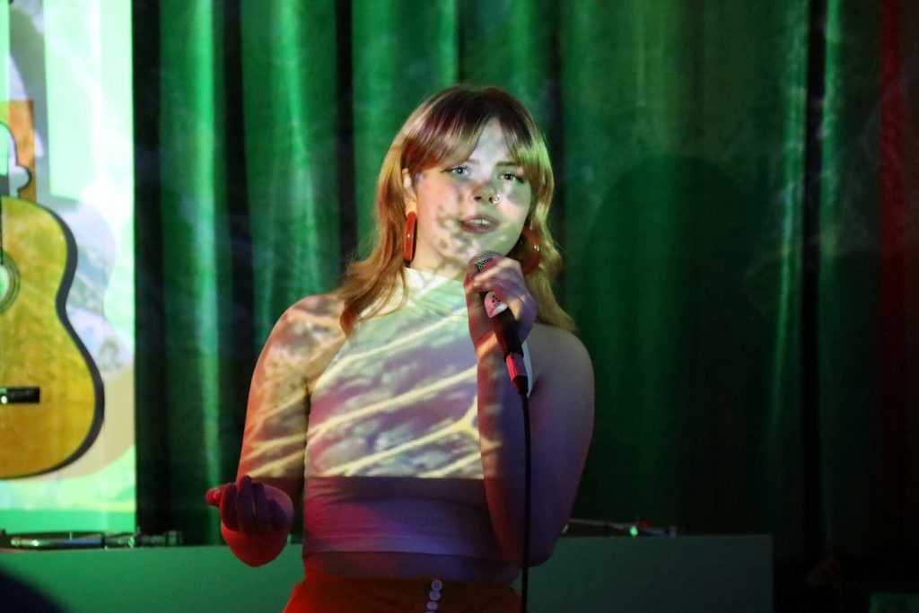 En ung kvinna sjunger i en mikrofon, med färgglada ljusmönster projicerade på hennes ansikte och kropp.