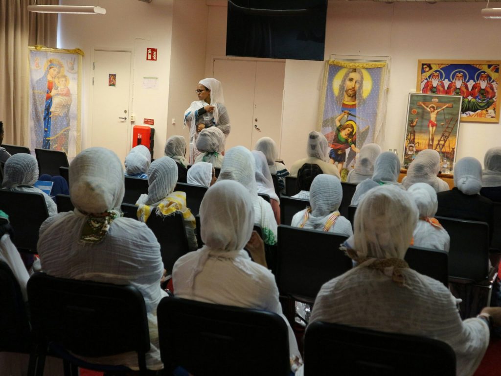 En grupp människor med vita huvudbonader deltar i en föreläsning i ett rum dekorerat med religiösa ikoner.