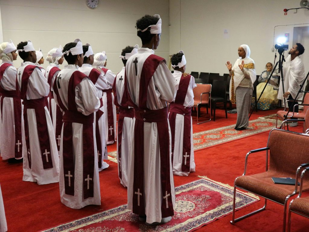 En grupp i vita dräkter med kors står i rader, vända mot en kvinna som talar i ett rum med röda mattor.