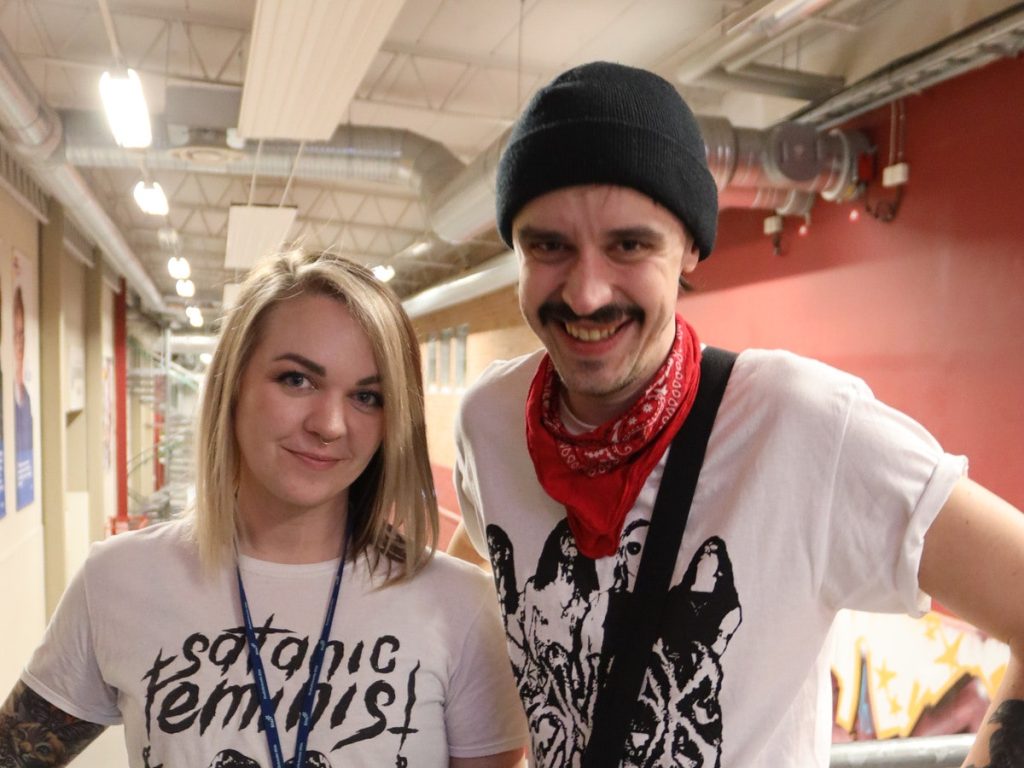 Två personer ler inomhus, den ena med blont hår och den andra med mustasch och svart mössa.