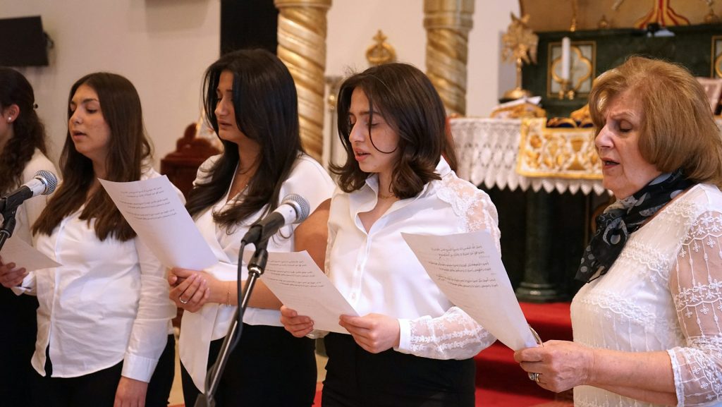 Fem kvinnor, klädda i vita toppar, sjunger med pappersark i händerna framför mikrofoner i en kyrka.