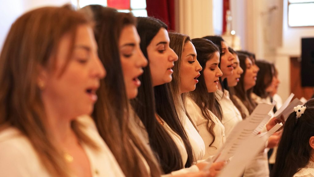 En grupp kvinnor i vita skjortor sjunger tillsammans inomhus, var och en med noter i handen.