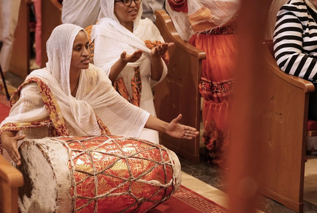 En kvinna i traditionell klädsel spelar på en stor trumma under en gudstjänst, medan andra ber i närheten.
