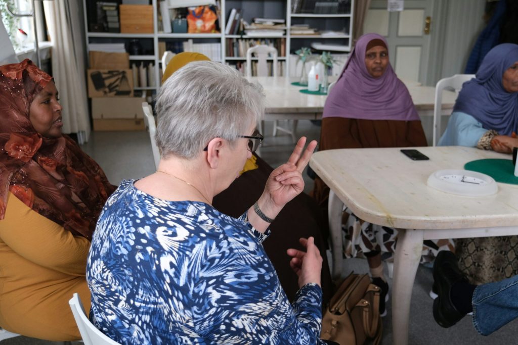 En kvinna med kort grått hår gör tecken med händerna medan andra lyssnar runt borden i en lokal.